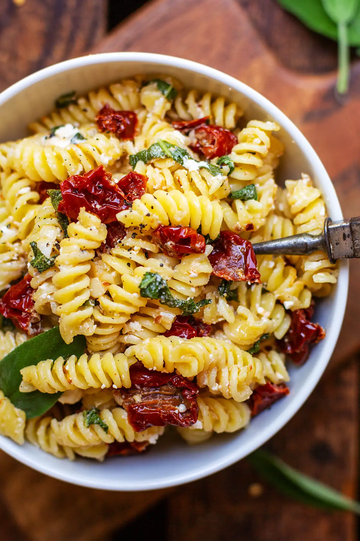 Fertige Pasta mit Salbei mit getrockneten Tomaten und Feta in einer weißen Schüssel auf dunklem Holzuntergrund. Garniert mit frischem Salbei und eine Gabel in der Schüssel - bereit zum Essen.
