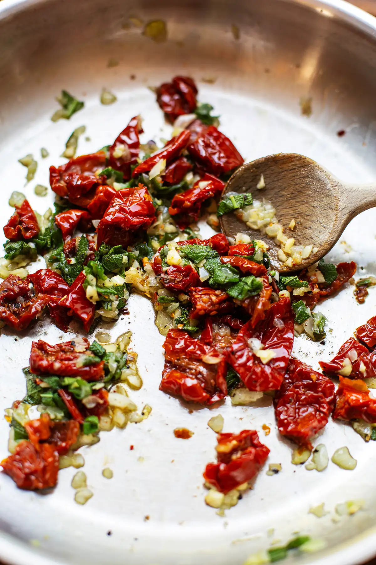 Klein geschnittene getrocknete Tomaten, Zwiebel, Knoblauch und Salbei werden mit einem Holzlöffel in einer Pfanne für die Salbei Pasta angebraten.