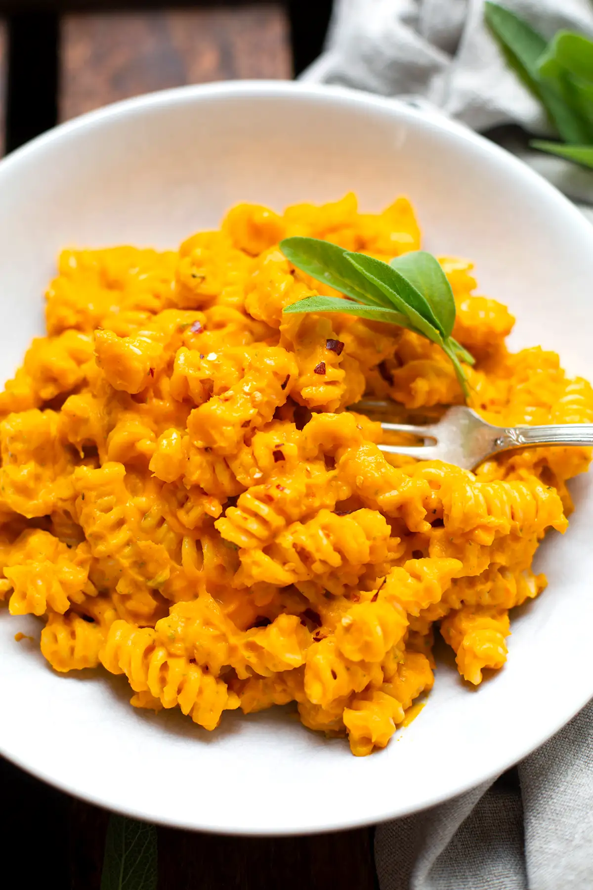 Fertige Kürbis Pasta mit Hokkaido und Salbei auf einem weißen Teller. Garniert mit frischem Salbei. In der Pasta steckt eine silberne Gabel. Der weiße Teller steht auf einem dunklen Holzuntergrund. 