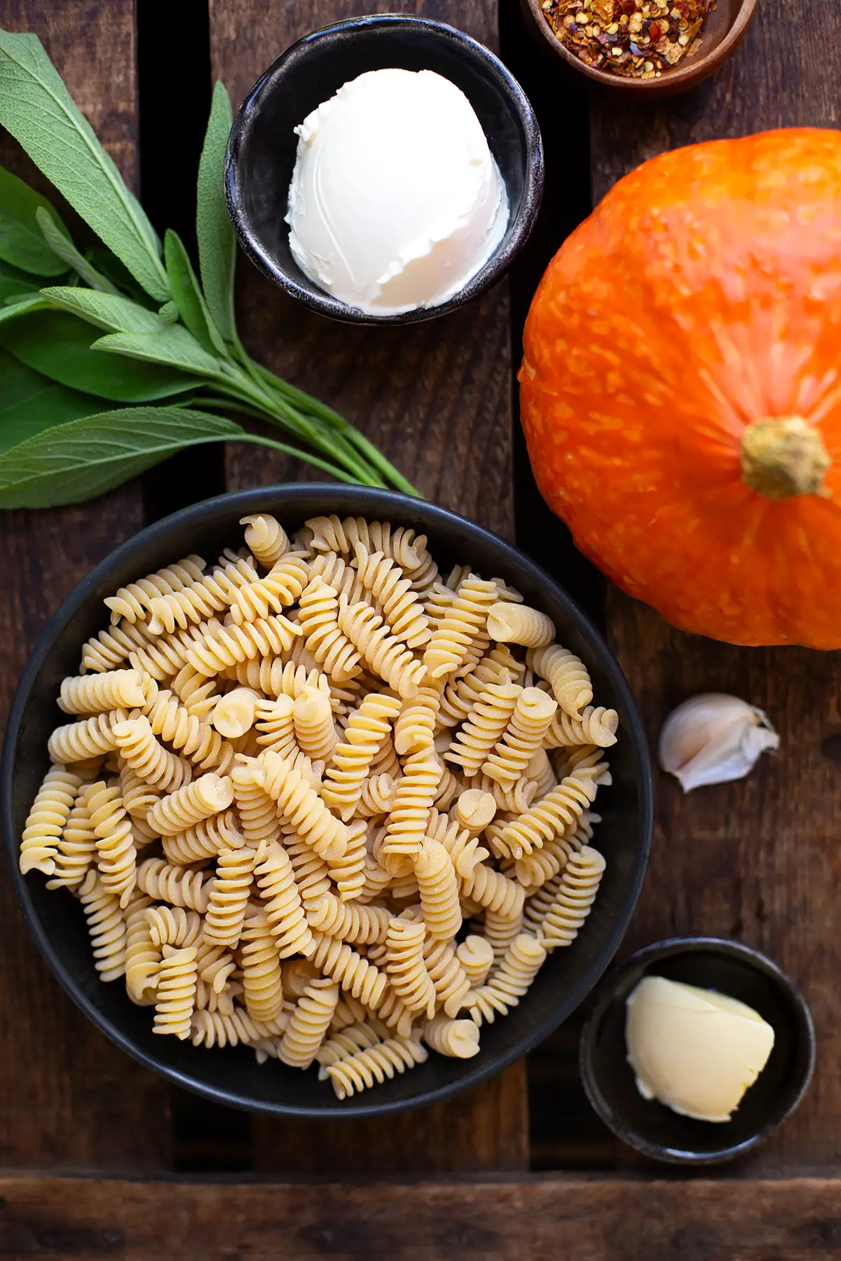 Einzelne Zutaten für die Pasta mit Kürbis auf dunklem Holzuntergrund. Zu sehen sind: Creme Fraiche, frischer Salbei, Hokkaidokürbis, Fusilli, Gewürze, Knoblauch und Butter.