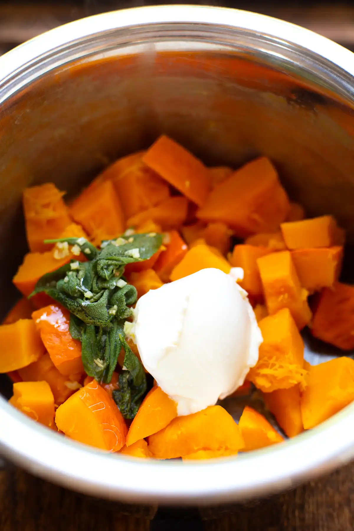 Klein geschnittener und gekochter Hokkaidokürbis zusammen mit Salbei, Knoblauch und Creme Fraiche in einem silbernen Topf kurz vor dem Pürieren.