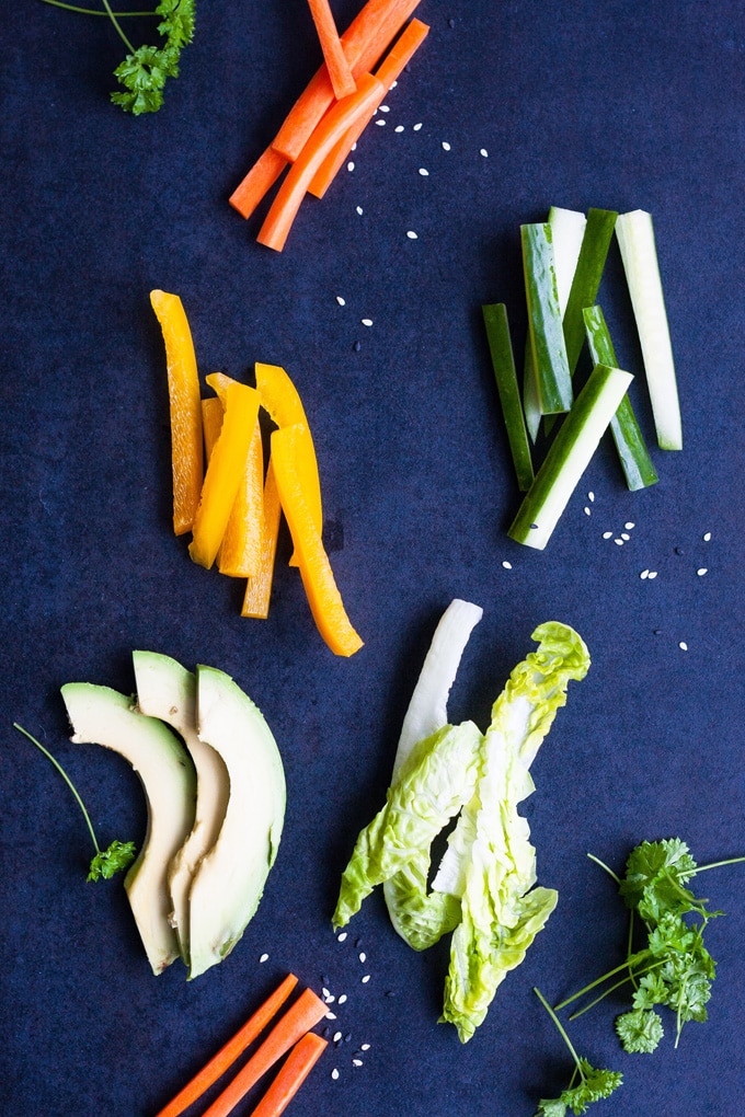 Werbung. Summer Rolls mit schneller Erdnusssauce. Frisch, knackig und gesund! Perfekt als leichtes Abendessen und zum Mitnehmen - kochkarussell.com Zutaten für die Sommerrollen in Streifen geschnitten, liegen auf einem blauen Untergrund. Zu sehen sind gelbe Paprika, Avocado, Gurken, Salat, Karotten.