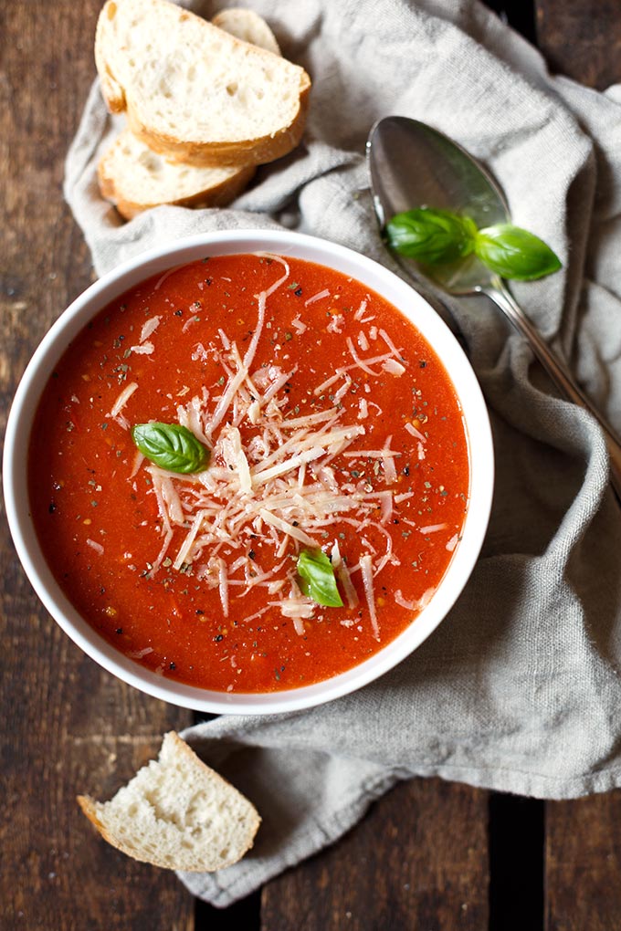 Fertige geröstete Tomatensuppe aus Tomaten, Knoblauch, Zwiebel, Olivenöl und Basilikum in einer weißen Schüssel. Daneben liegen zwei Scheiben Baguette, ein silberner Löffel und ein graues Geschirrhandtuch.