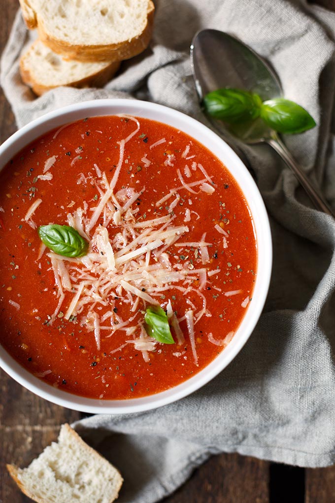 Fertige geröstete Tomatensuppe  aus dem Ofen mit Parmesan und Basilikum garniert. Neben der weißen Schüssel liegt ein silberner Löffel, Basilikum und ein graues Geschirrtuch.