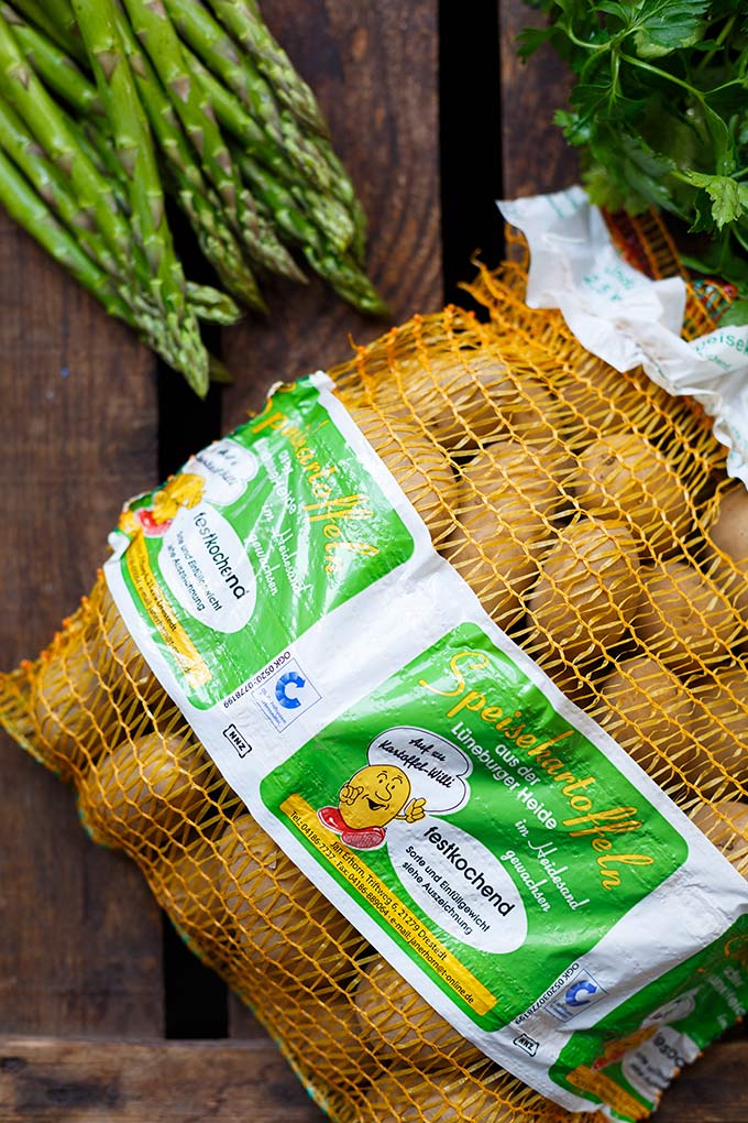 Ein Sack Kartoffeln auf dunklem Holzuntergrund. Daneben liegen ein paar Stangen grüner Spargel und Petersilie.