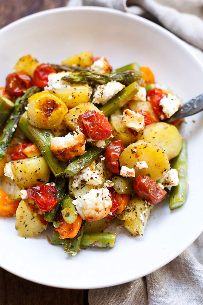 Fertige Ofenkartoffeln mit grünem Spargel, Tomaten und Feta auf einem weißen Teller.