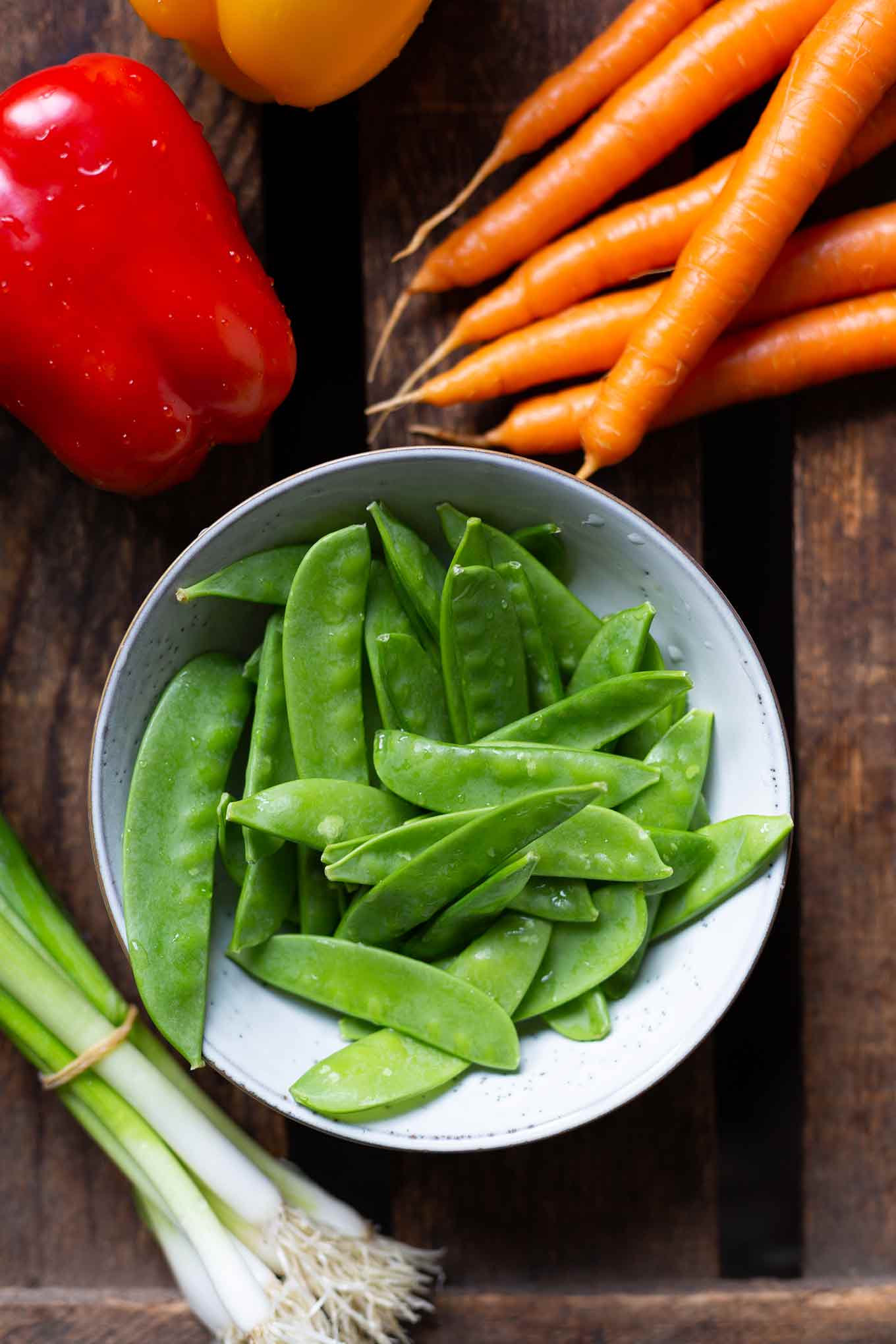 Einzelne Zutaten für den asiatischen Spaghettisalat. Zu sehen sind eine rote Paprika, Möhren, Zuckerschoten, Frühlingszwiebeln und eine gelbe Paprika. 