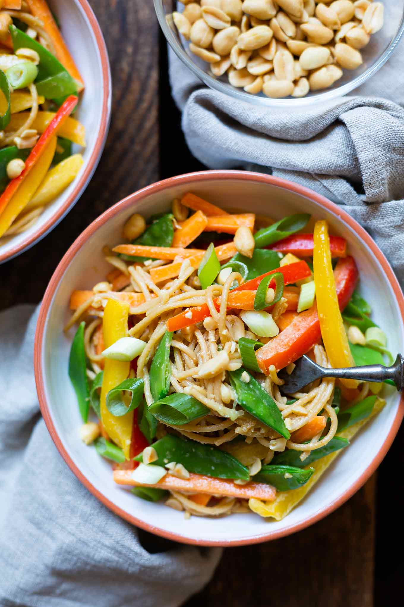Fertiger asiatischer Nudelsalat mit Erdnusssauce aufgeteilt in zwei weißen Schüsseln mit orangem Rand. Im Hintergrund eine weitere Schüssel mit Erdnüssen. Die Schüsseln stehen auf einem dunklen Holzuntergrund und einem grau-blauen Geschirrtuch.