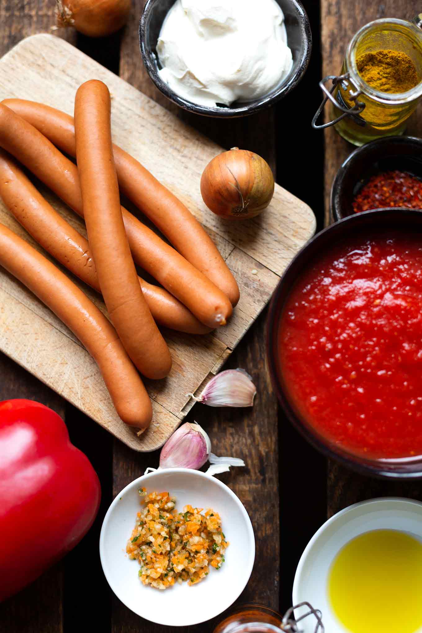 Die Zutaten für das Wurtgulasch: Wiener Würstchen und eine Zwiebel auf einem Holzbrett. Daneben eine Knoblauchzehe. In Schüsseln gehackte Tomaten, Gewürze, Currypulver und Creme Fraiche.