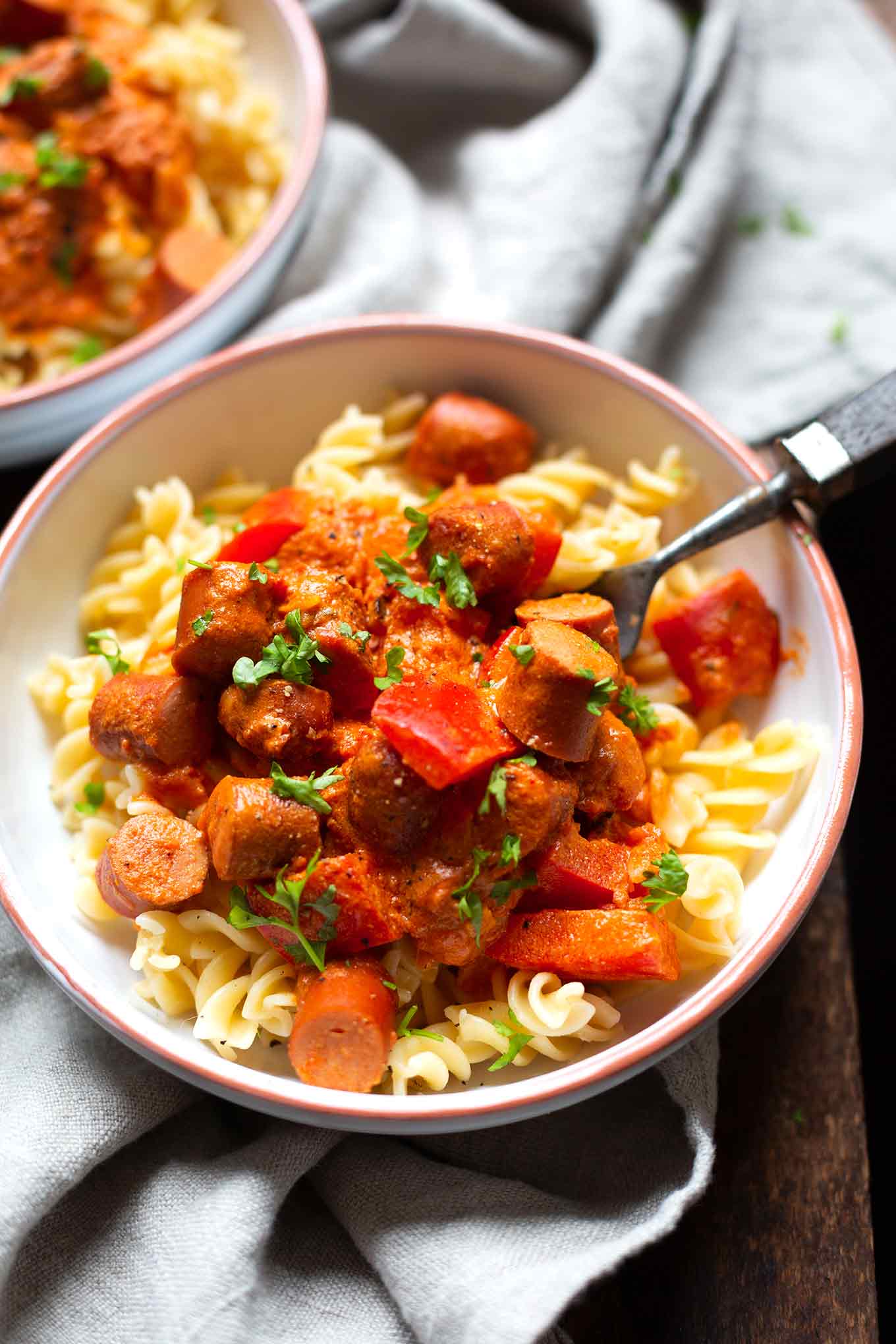 Fertiges Wurstgulasch in einer weißen Schüssel mit Fusili und einer Gabel. Im Hintergrund ist eine zweite Schüssel mit Gulasch zu sehen.
