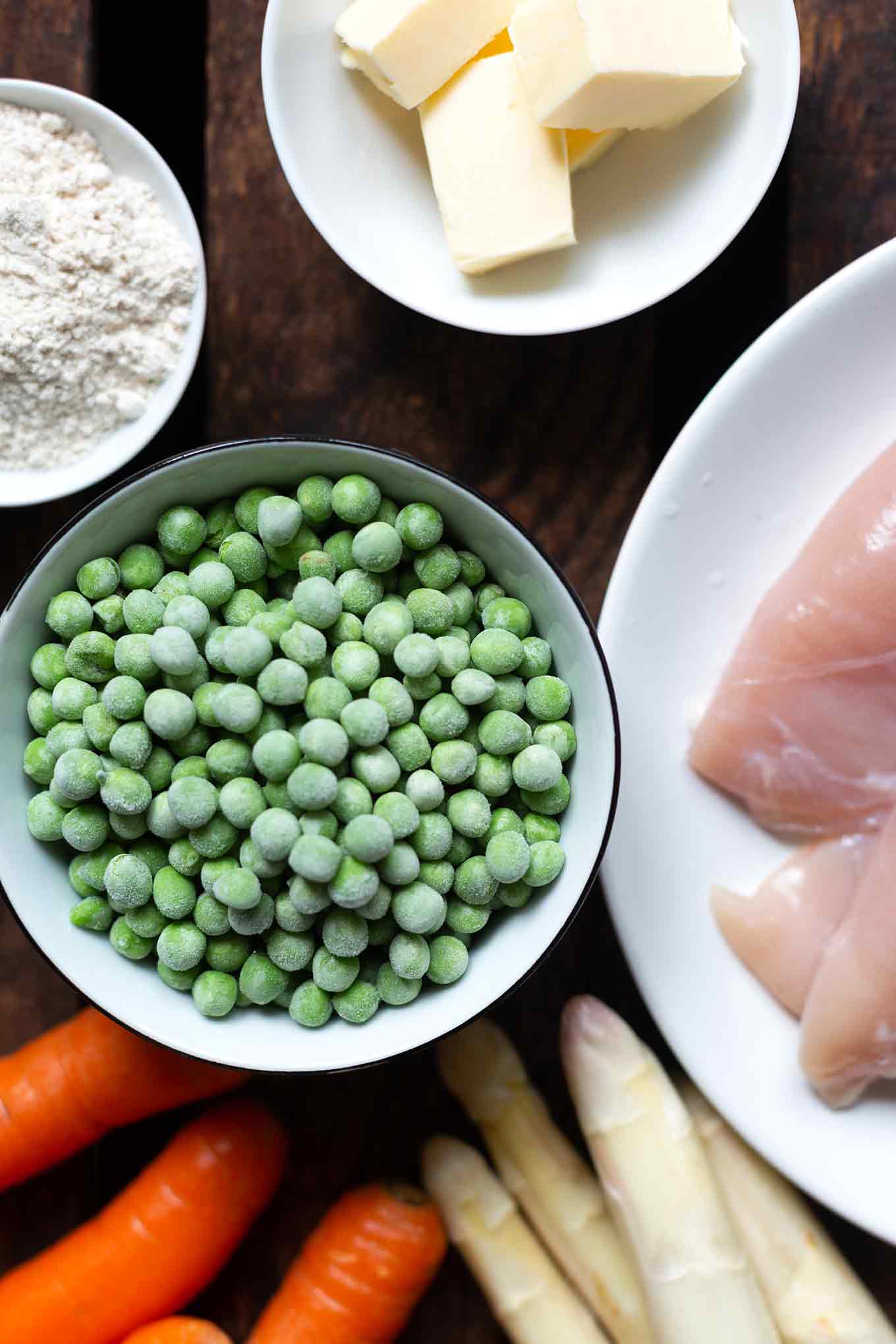 Zutaten für das einfache Hühnerfrikassee einzeln präsentiert. Zu sehen sind weißer Spargel, Karotten, Erbsen, Hühnerbrust, Mehl & Butter.