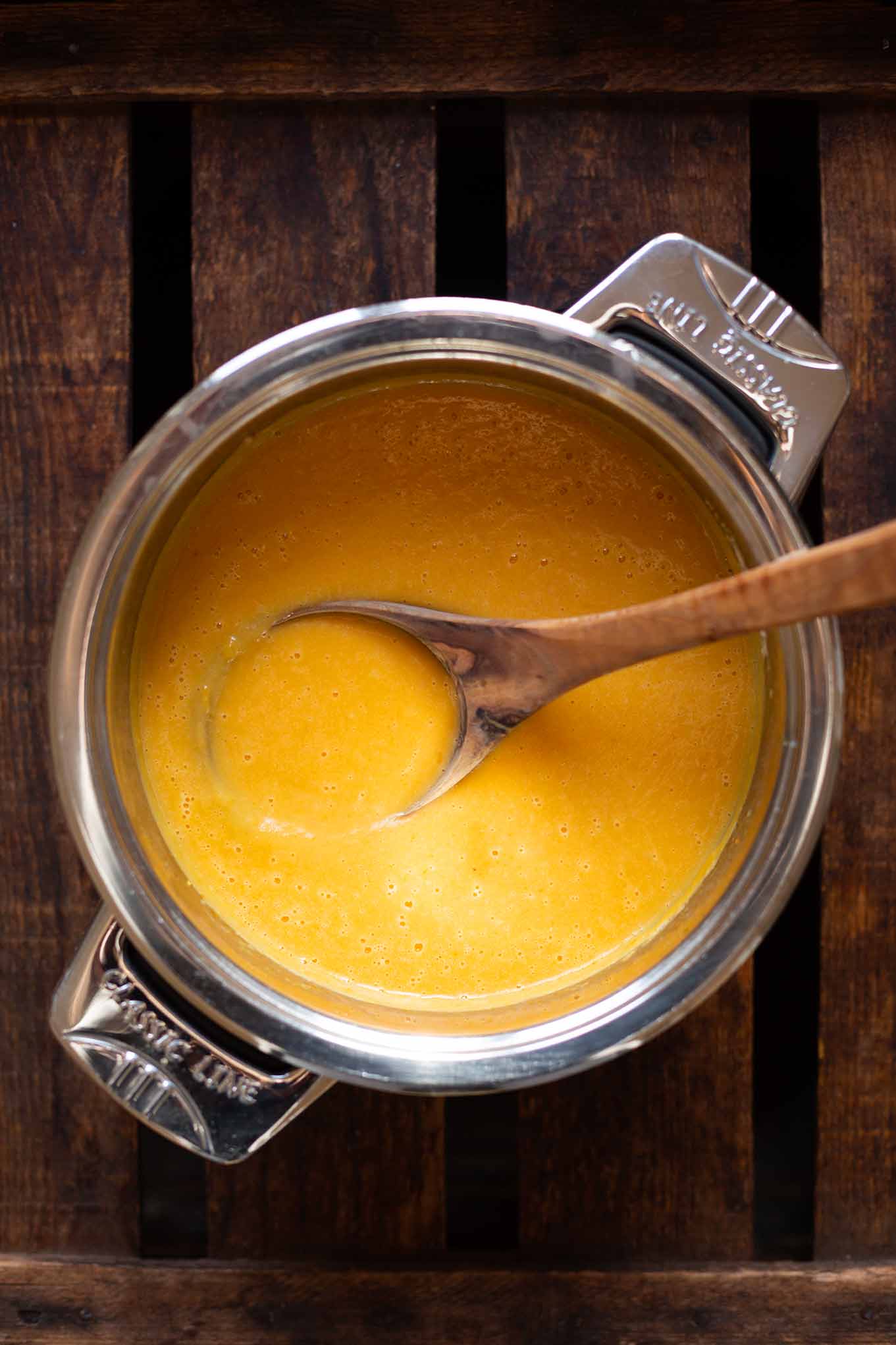 Pürierte vegane Kürbissuppe in einem silbernen Topf mit Holz-Kochlöffel. Der Topf steht auf einem dunklen Holzuntergrund.
