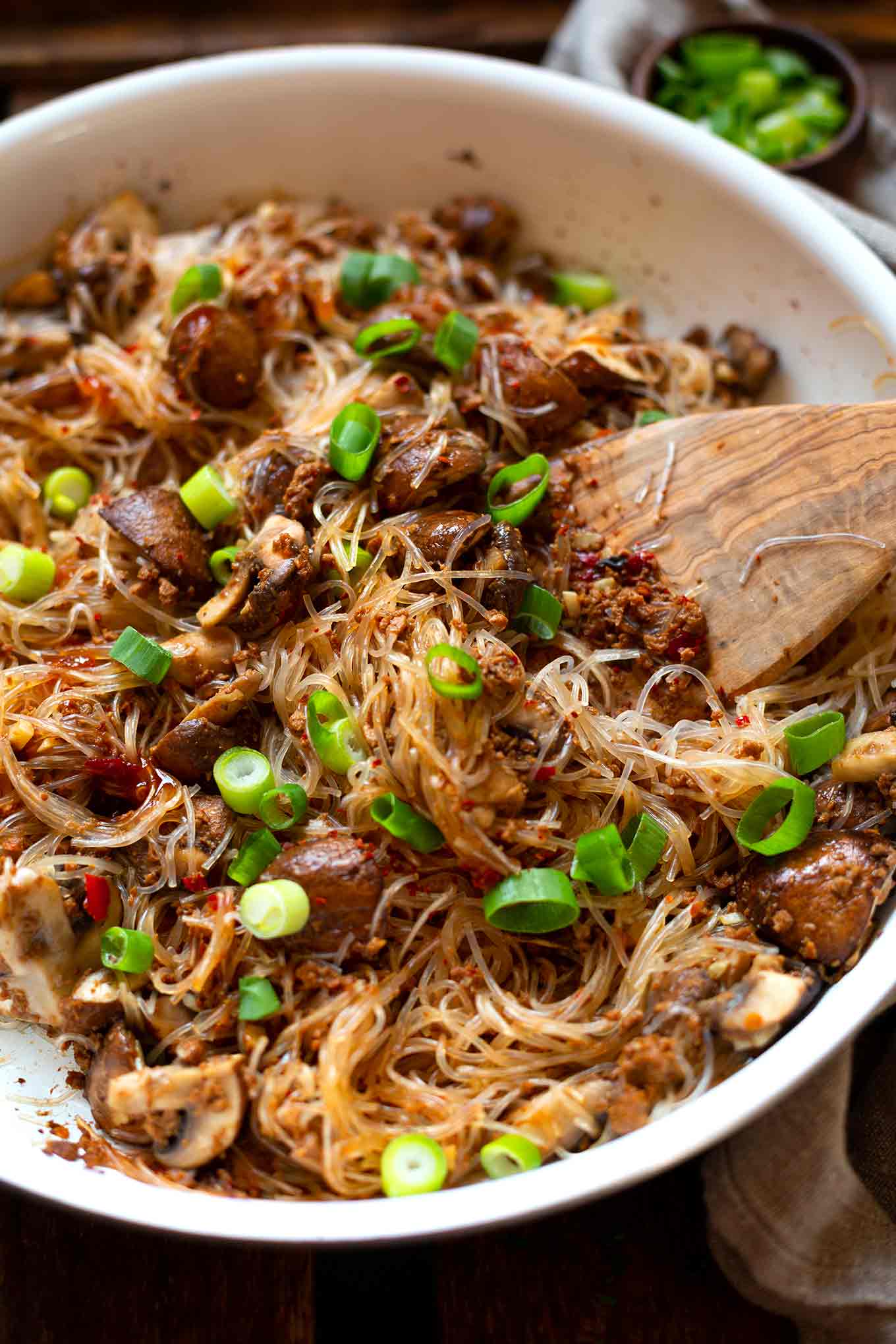 Nahaufnahme der fertigen asiatischen Glasnudel-Hackfleisch-Pfanne angerichtet in einer Schüssel mit Frühlingszwiebeln und einem großen Holzlöffel zum Servieren. Im Hintergrund frische, geschnittene Frühlingszwiebel in einer kleinen Schüssel.