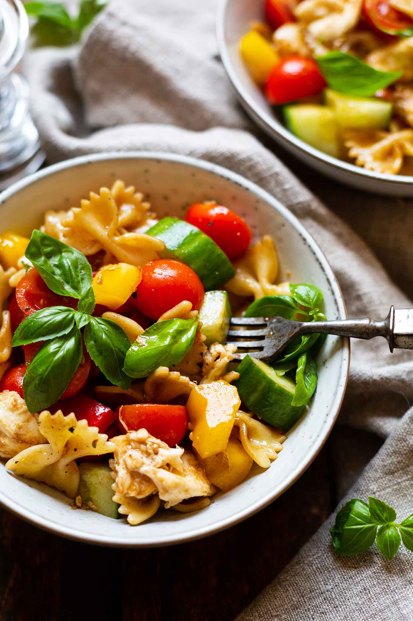 Bunter Nudelsalat aufgeteilt in zwei Schüsseln, garniert mit Basilikum. In der vorderen Schüssel mit einer Gabel.