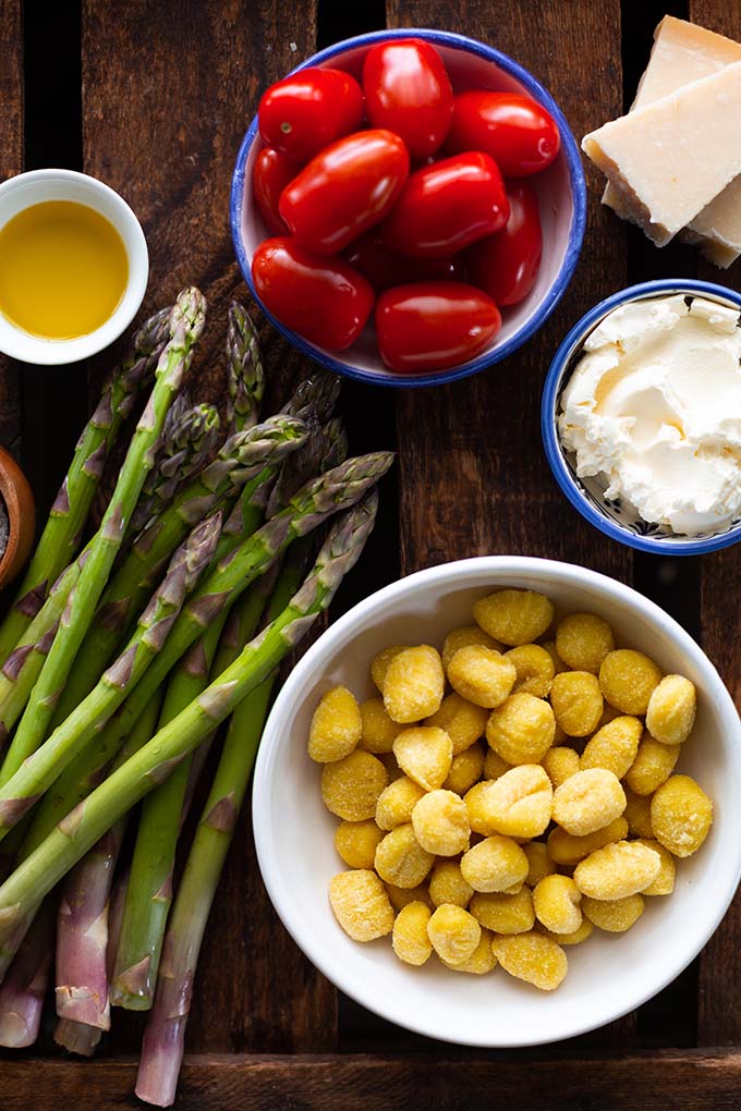 Zutaten für grüner Spargel mit Gnocchi und Tomaten: Grüner Spargel, Gnocchi, Tomaten, Creme Fraiche, Olivenöl und Parmesan auf einem Holzuntergrund.