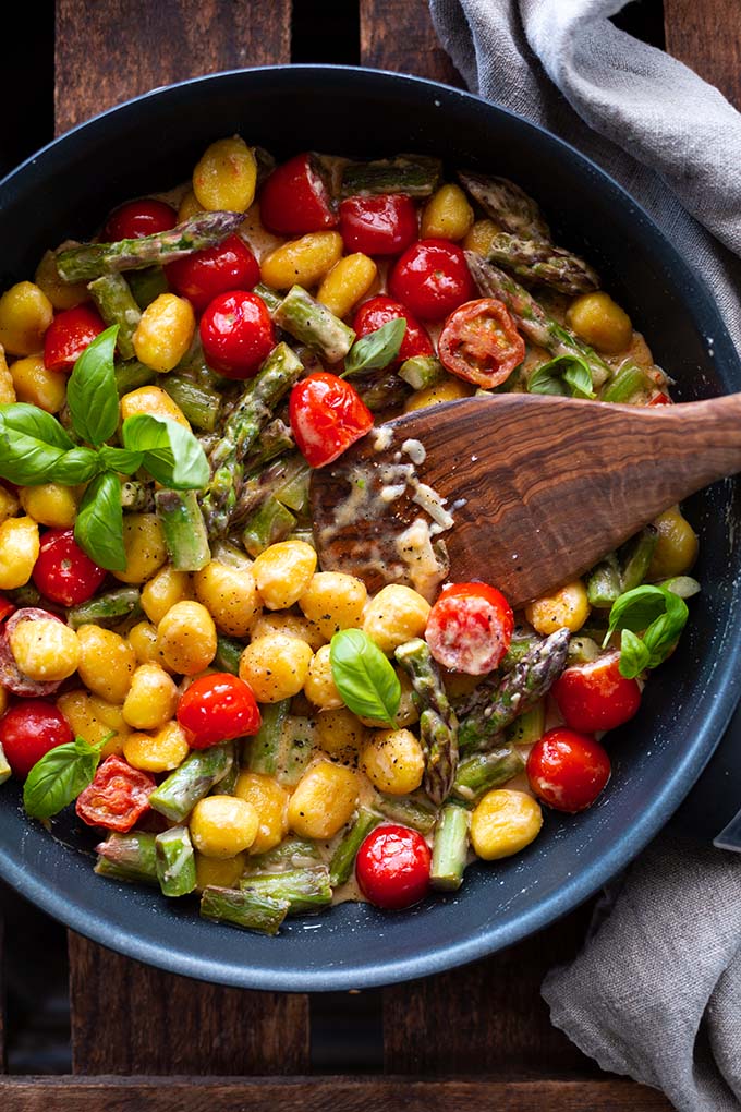 Grüner Spargel gebraten in der Pfanne mit Gnocchi, Tomaten und cremiger Sauce von oben.
Mit einem Pfannenwender aus Holz in der Pfanne und einem grau-blauen Geschirrtuch im Hintergrund.