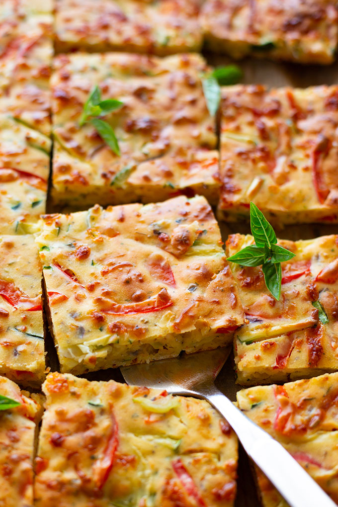 Fertige Gemüse-Quiche auf einem Backblech und in Quadrate geschnitten. Ein Stück wird mit einem Tortenheber leicht angehoben.