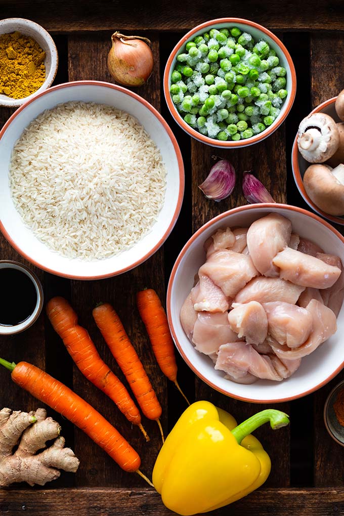 Zutaten für das Nasi Goreng in kleinen Schüsseln und auf einem dunklen Untergrund. Wir sehen Kurkuma, Erbsen, Paprika, Zwiebeln, Pilze, Knoblauch, Soja Sauce, Hähnchenfleisch und Karotten.