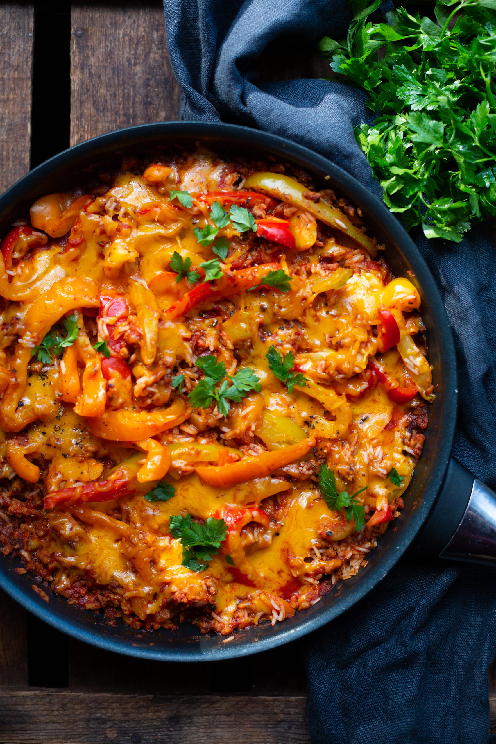 Rezeptfotografie vegetarische Paprika-Reispfanne mit Hackfleisch. Abgebildet in einer Pfanne auf einem dunklen Hintergrund und garniert mit Petersilie.