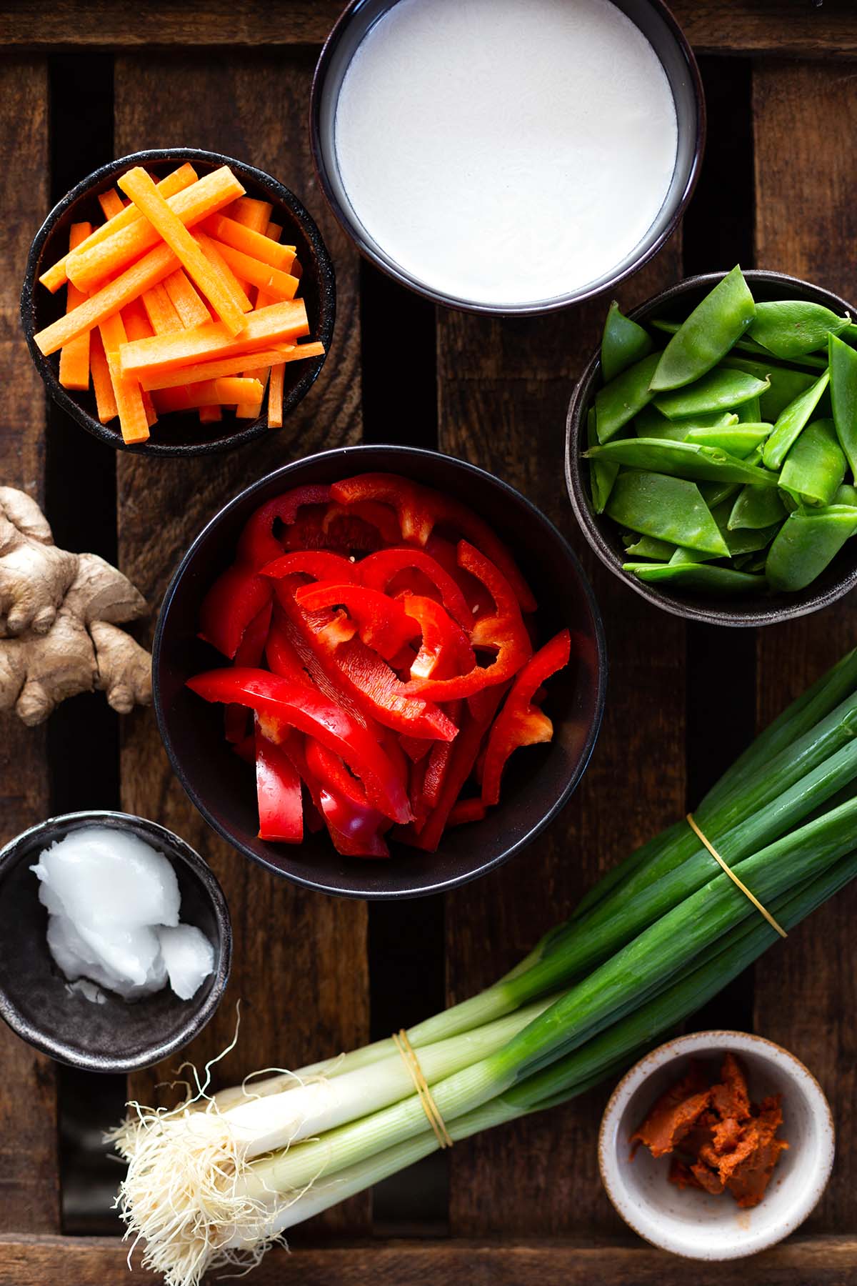 Zutaten für ein veganes, rotes Thai-Curry mit Kokosmilch in verschiedenen Schüsseln abgebildet. Wir sehen geschnittene Paprika, Kokosöl und Kokosmilch, Ingwer, ein Bund Lauchzwiebeln, rote Currypaste, Möhrenstifte und Zuckerschoten.