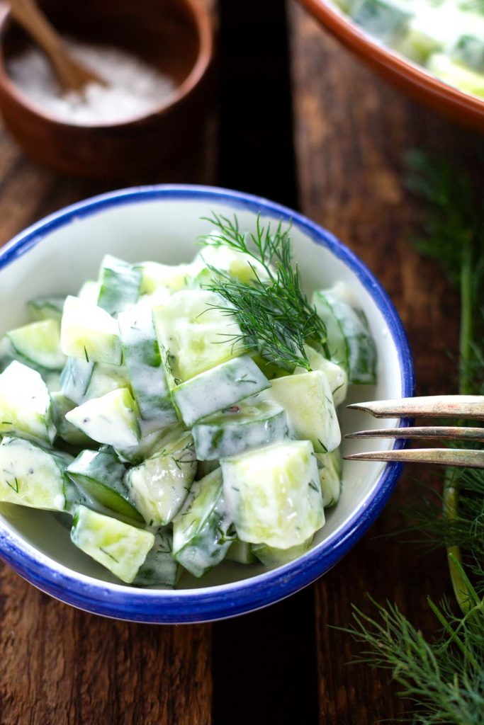 Gurkensalat mit Joghurt-Dressing und Dill in einer kleinen weißen Schüssel mit blauem Rand. Eine silberne Gabel lehnt an dem Rand der Schüssel.