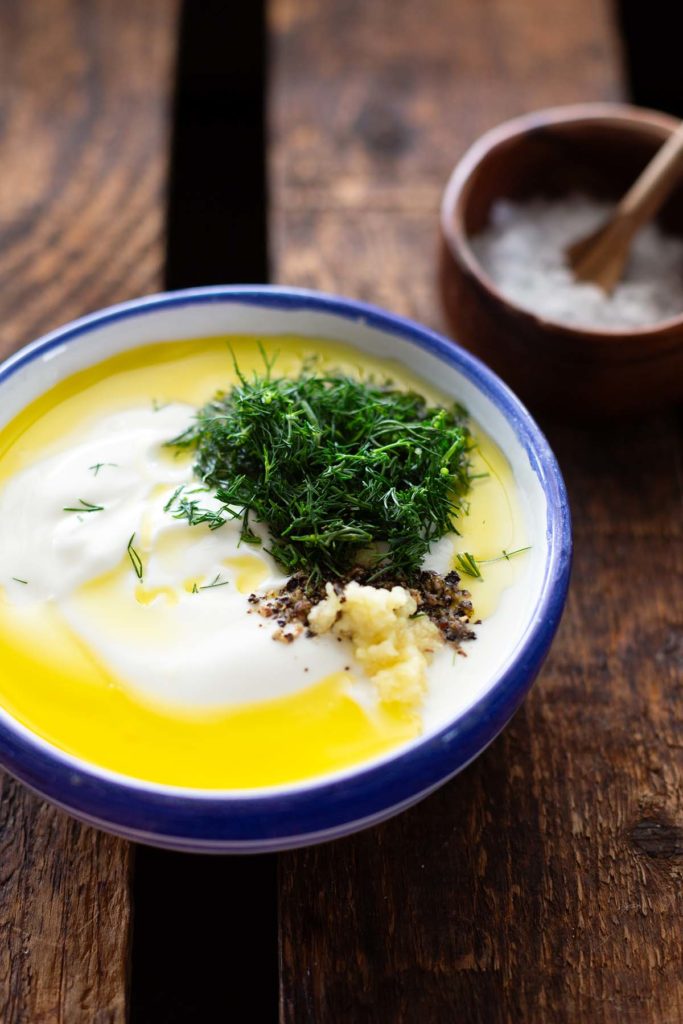 Gurkensalat-Dressing aus Joghurt, Dill, Öl, Zitronensaft, Knoblauch, Salz und Pfeffer in einer weißen Schüssel mit blauem Rand. Die Zutaten sind noch nicht vermischt. Die Schüssel steht auf einem dunklen Holzuntergrund und im Hintergrund ist eine weitere kleine Holzschüssel mit Salz und einem Holzlöffel zu sehen.