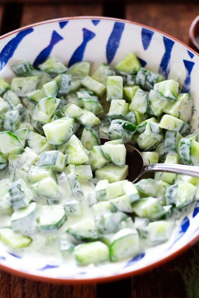 Fertiger Gurkensalat mit Dill in einer großen weißen Schüssel mit braunem Rand und blauen Fischen. Die Schüssel steht auf einem dunklen Holzuntergrund. Im Gurkensalat steckt ein silberner Löffel - bereit zum Servieren.