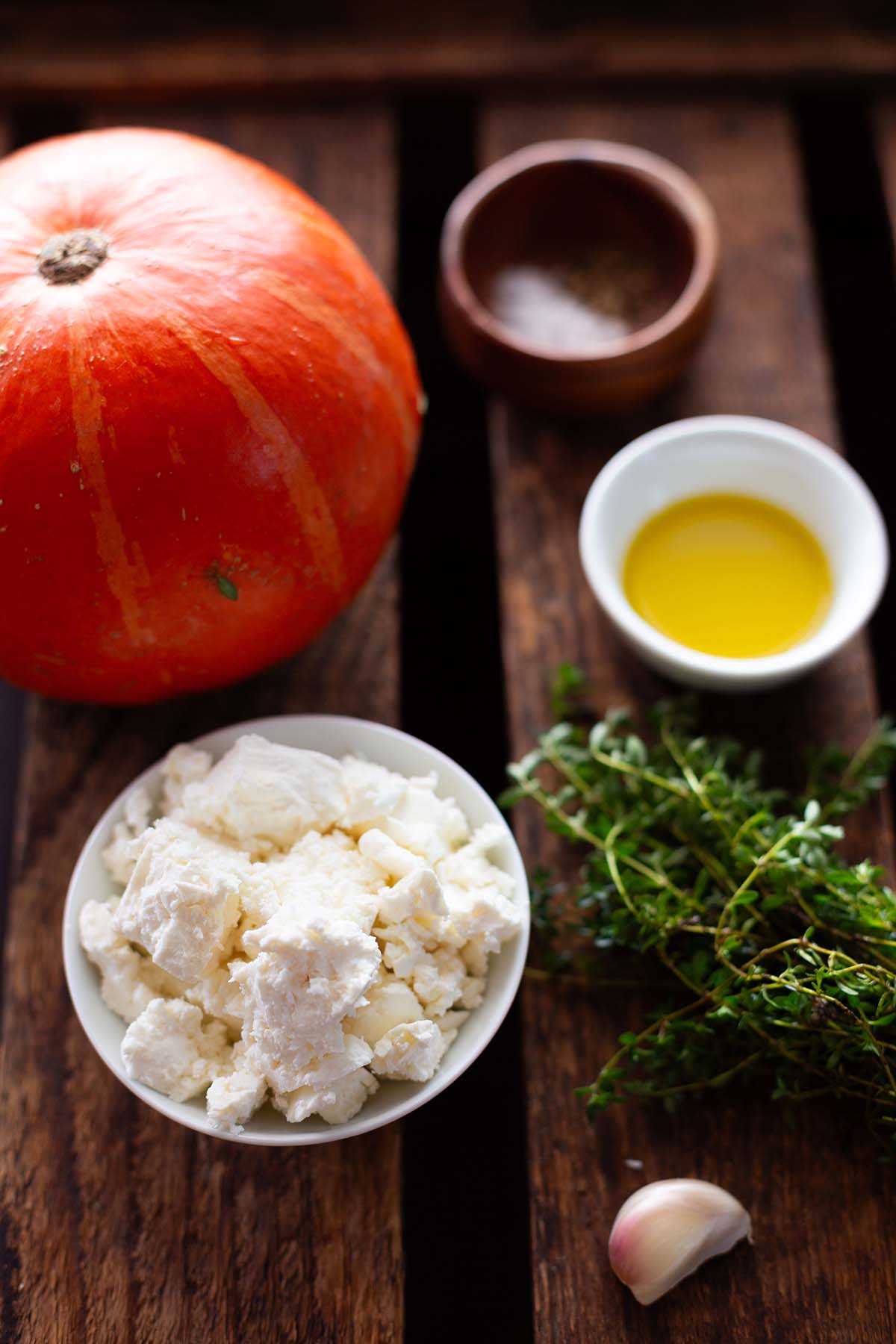 Zutaten für den Kürbis aus dem Ofen mit Feta auf einem braunen Untergrund. Es sind frischer Thymian, Feta, Olivenöl und ein Hokkaido-Kürbis zu sehen.