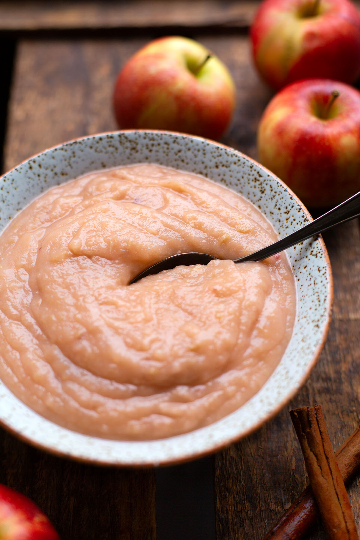 Selbst gemachtes Apfelmus in einer gesprenkelten Steingut Schüssel mit einen Löffel garniert, auf einem dunklen Holzuntergrund angerichtet und mit Äpfeln im Hintergrund.