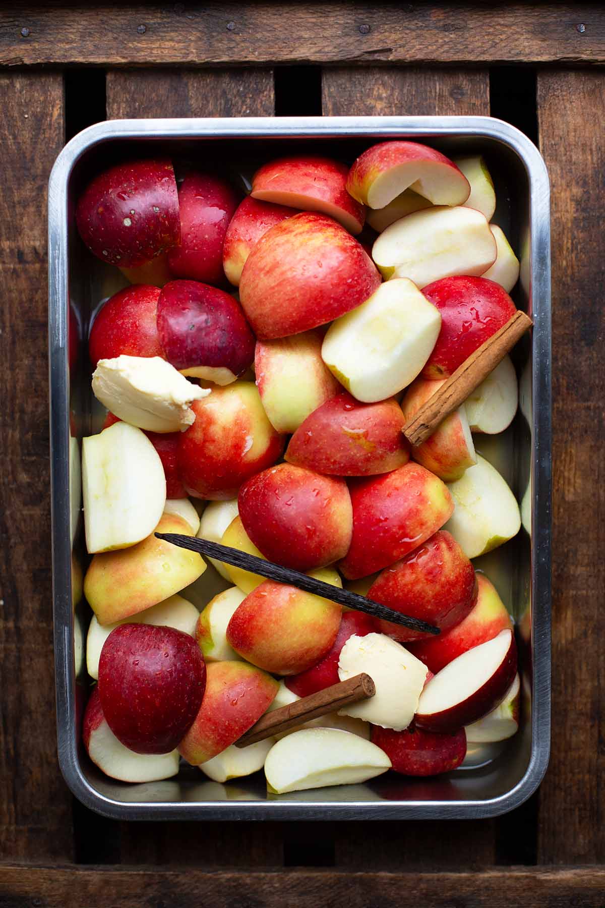 Apfelmus selber machen im Ofen. Dazu wurden alle klein geschnitten Äpfel mit Zimtsangen, Vanilleschote, Butter und Apfelsaft in eine große Auflaufform gegeben.Auf einem dunklen Holzuntergrund als Flatlay fotografiert.