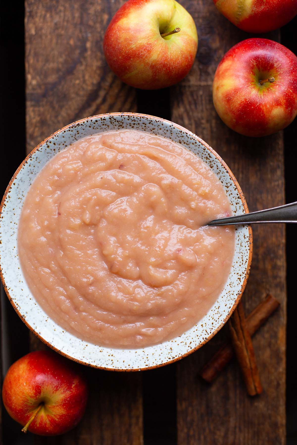Selbst gemachtes Apfelmus in einer gesprenkelten Steingut Schüssel mit einen Löffel garniert, auf einem dunklen Holzuntergrund drapiert und mit Äpfeln im Hintergrund. Als Flatlay von oben fotografiert.