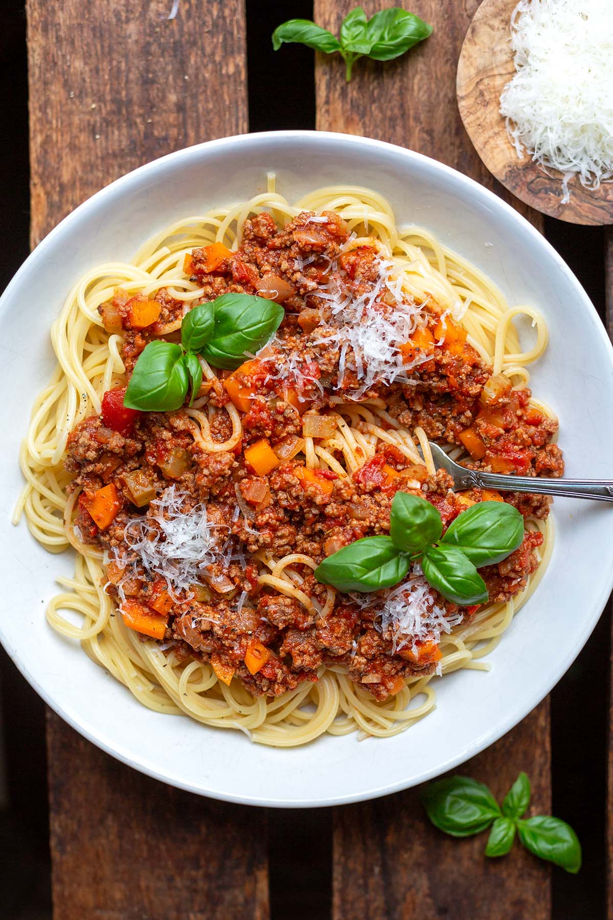 Spaghetti mit Bolognese Sauce angerichtet auf einem weißen Teller angerichtet mit Parmesan und Basilikum auf einem dunklen Holzuntergrund.