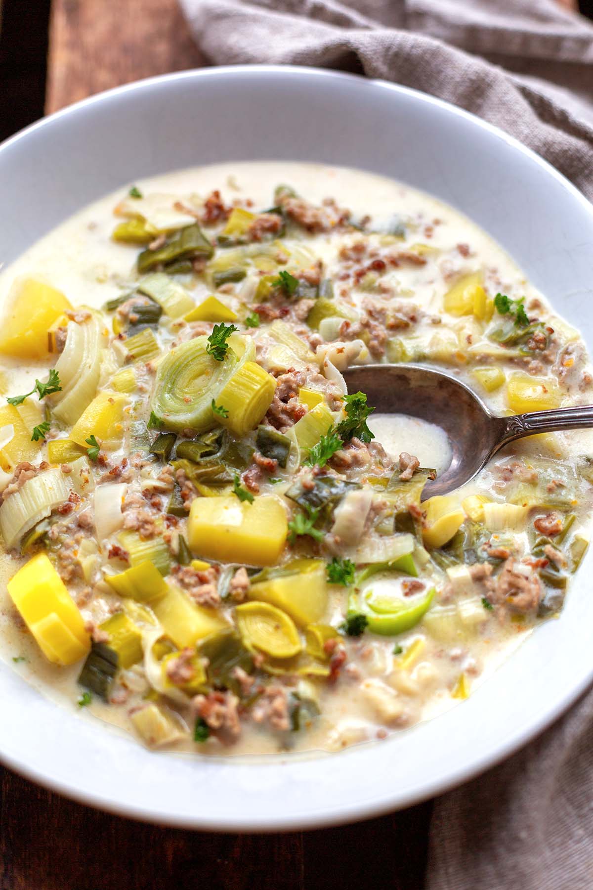 Käse-Lauch-Suppe iim Close-up in einem weißen Teller auf einem dunklen Holzuntergrund mit einem beige-grauen Leinentuch und antikem Löffel dekoriert.