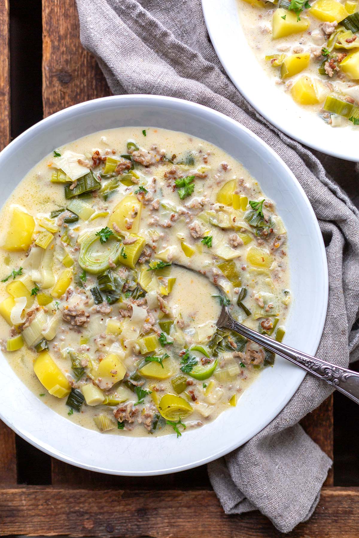 Käse-Lauch-Suppe in einem weißen Teller auf einem dunklen Holzuntergrund mit einem beige-grauen Leinentuch dekoriert.