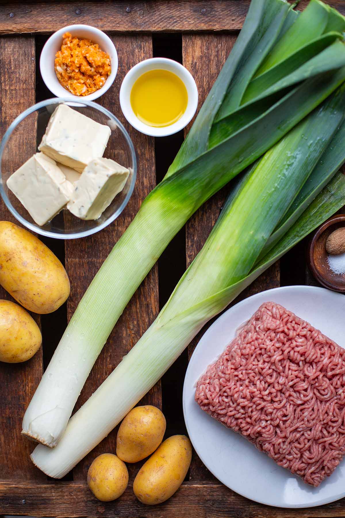 Die Zutaten für die Hack-Lauch-Suppe auf einem dunklen Holzuntergrund. Zu sehen sind zwei Stangen Lauch, Kartoffeln, Brühe, Olivenöl, Hackfleisch, Kräuter-Schmelzkäse und Gewürze.