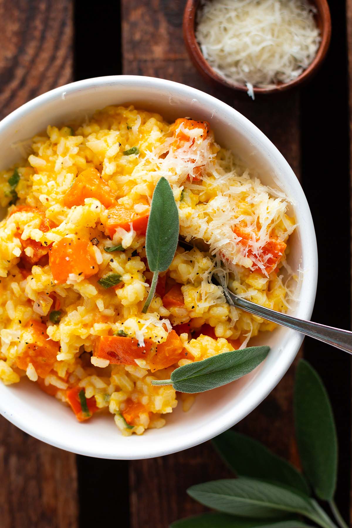 Kürbisrisotto mit Salbei & Parmesan in einer weißen Schüssel auf einem dunklen Holzhintergrund. Garniert mit Salbeiblättern.