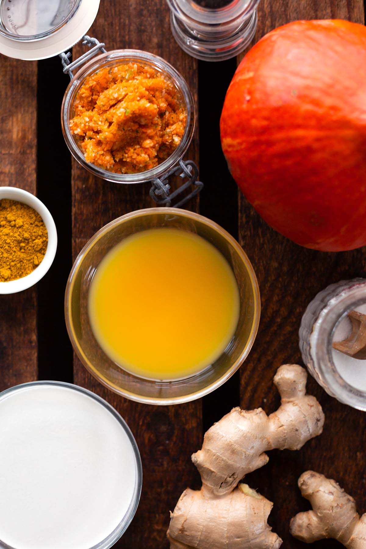 Zutaten für die Kürbissuppe mit Kokosmilch und Ingwer: Zu sehen sind ein ganzer Hokkaidokürbis, Ingwer, Kokosmilch, Gemüsebrühe, Currypulver, Salz und Pfeffer in einzelnen Schälchen.