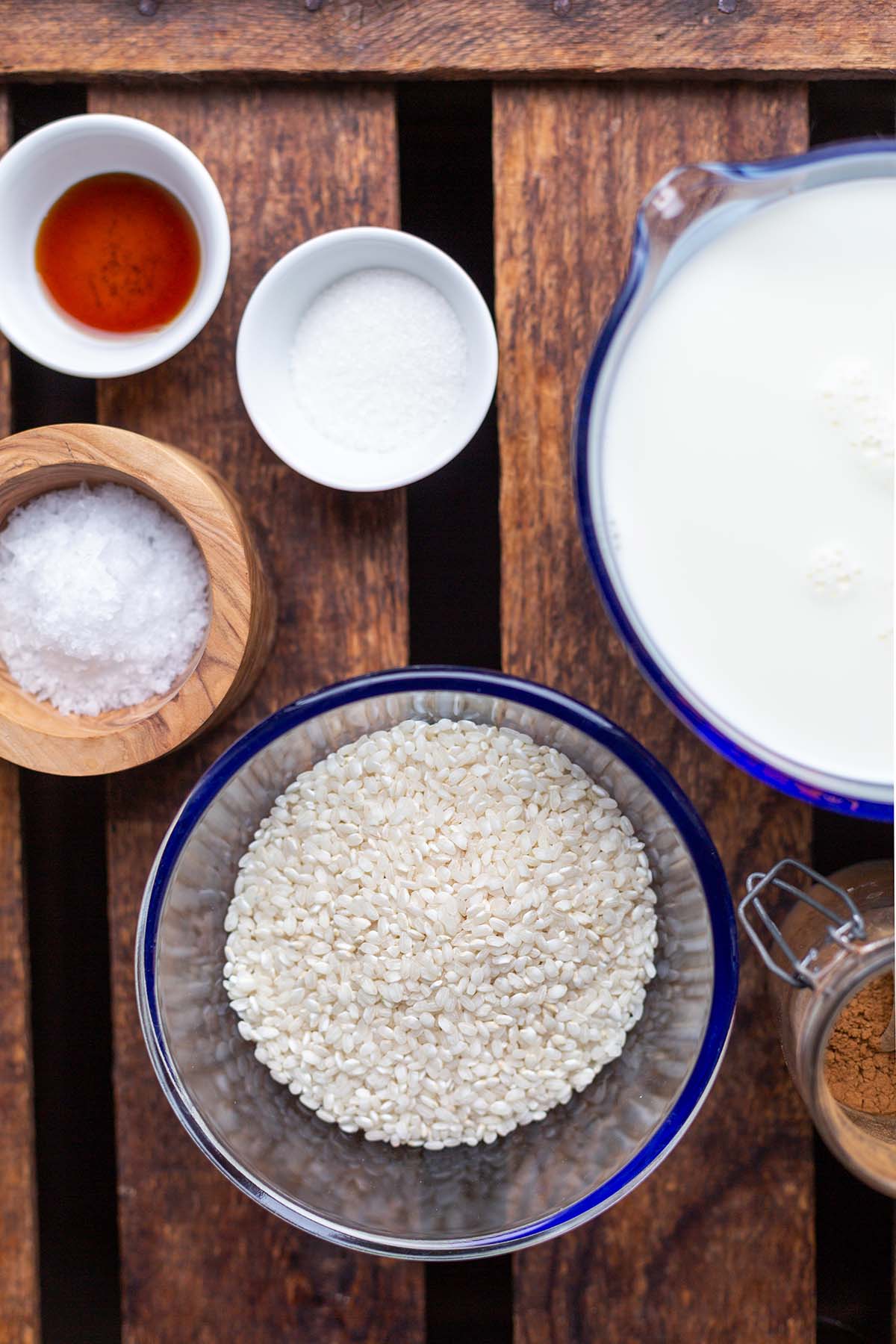 Alle Zutaten für de Milchreis in kleinen Schüsseln auf einem dunklen Holzuntergrund. Zu sehen sind Milch, Zucker, Rundkornreis, Zimt & Vanilleextrakt.