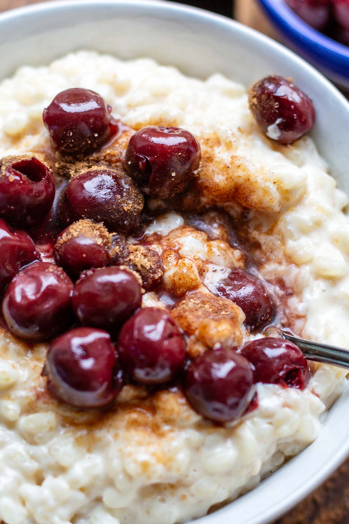Milchreis im Close-up angerichtet mit heißen Kirschen und Zimt&Zucker in einer weißen Schüssel auf einem dunklen Holzuntergrund.