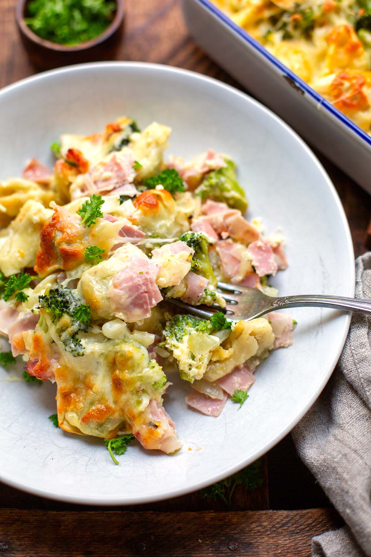 Tortellini-Auflauf mit Schinken und Brokkoli  auf einem weißen Teller portionsweise angerichtet auf einem dunklen Holzuntergrund mit einem grauen Küchentuch drapiert.