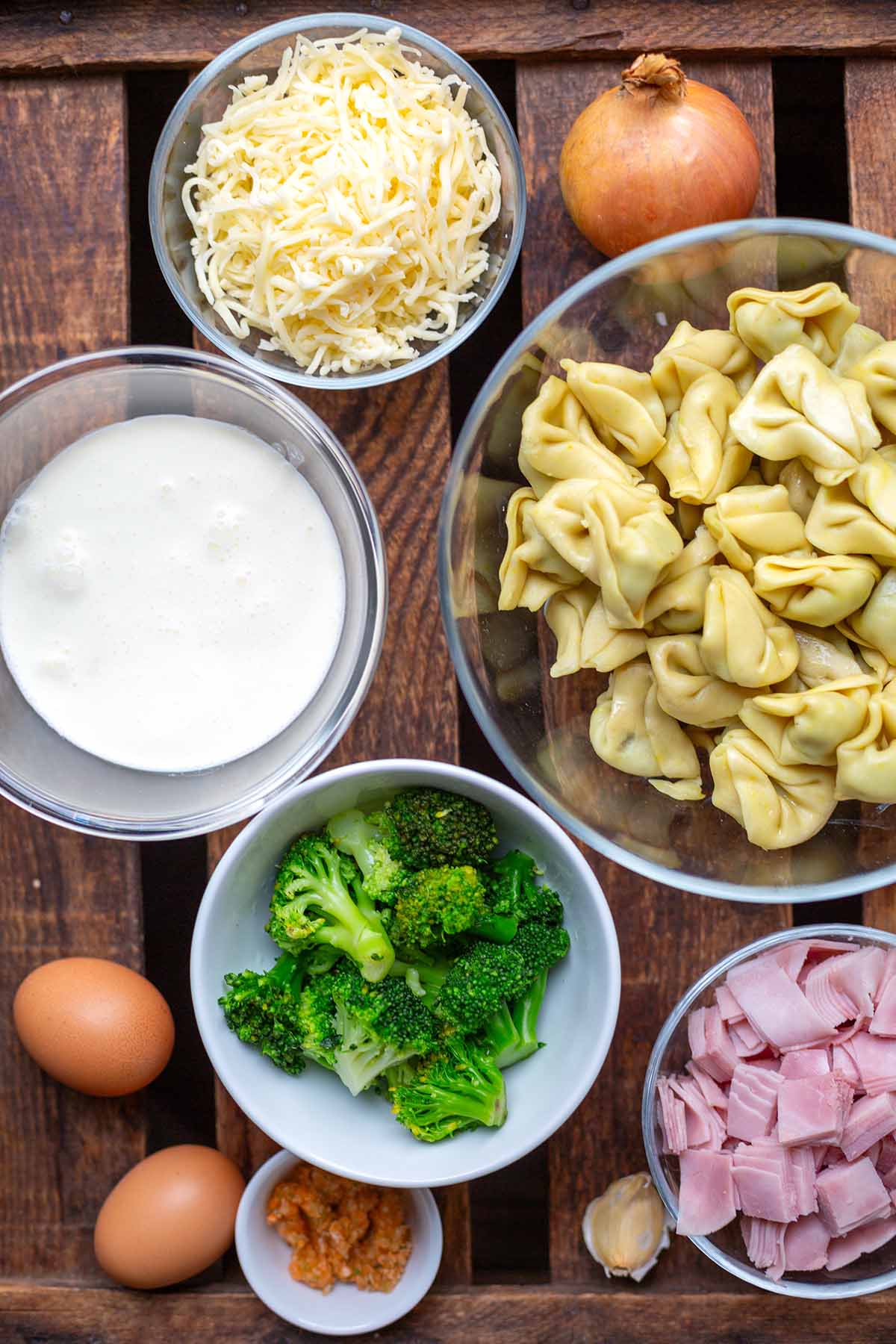 Die Zutaten für den Tortellini-Auflauf in verschiedenen Schüsseln verteilt auf einem dunklen Holzuntergrund. Zu sehen sind Käse, frische Tortellini, gedünsteter Brokkoli, Eier, Sahne, Schinken, Gemüsebrühe + Knoblauch.