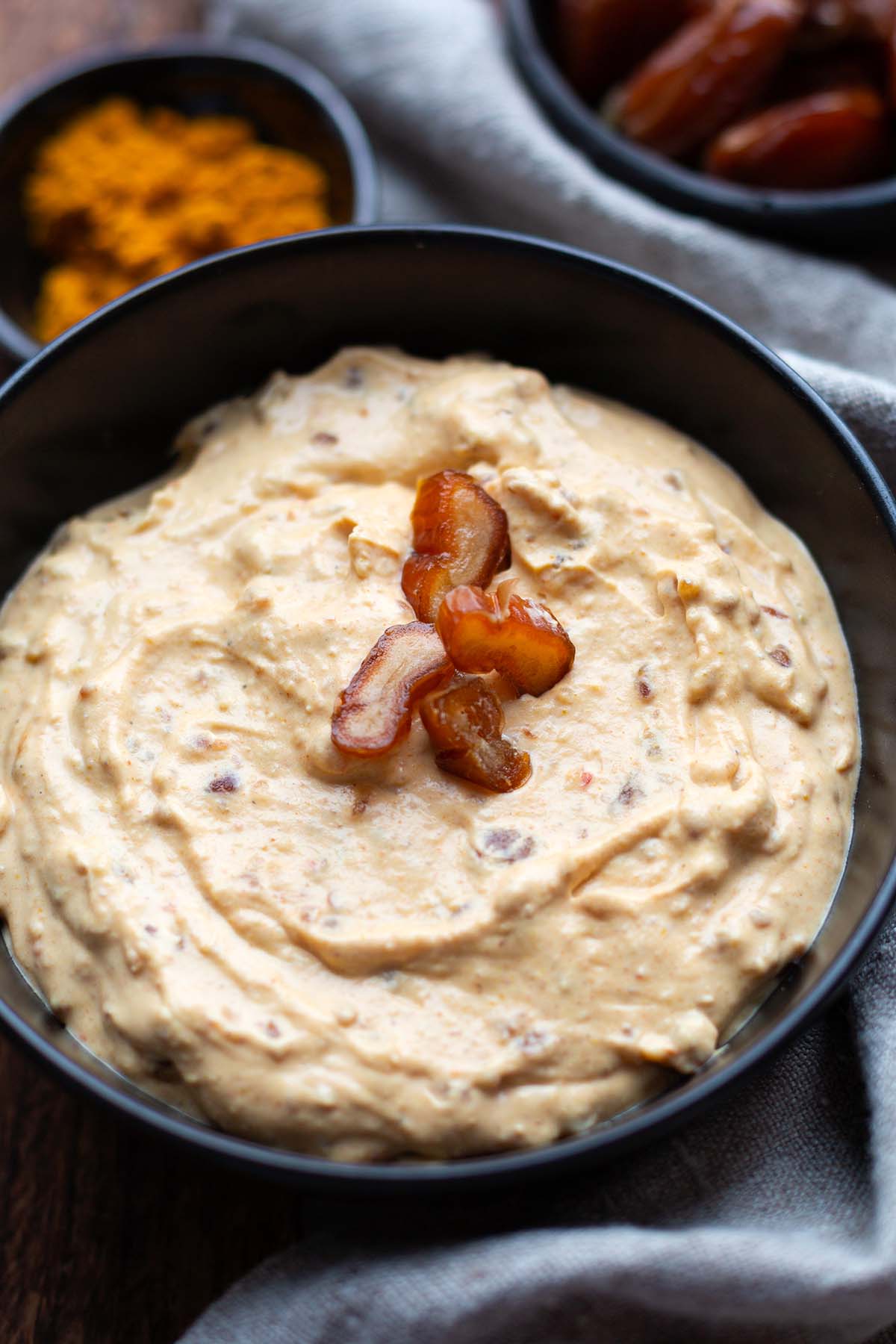Datteldip mit Curry und Frischkäse in einer schwarzen Schüssel. Currypulver und Datteln im Hintergrund.