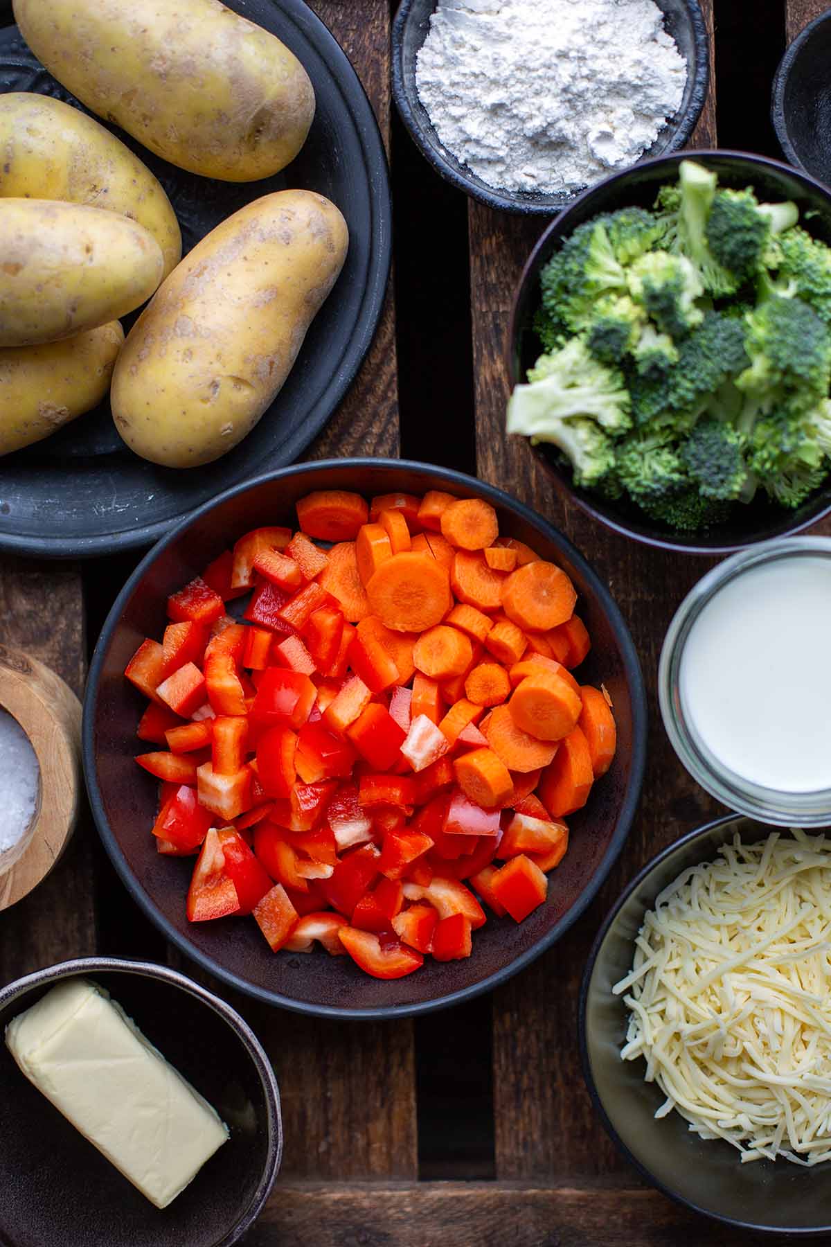 Die Zutaten für den Gemüseauflauf. Zu sehen sind rohe, ungeschälte Kartoffeln, Mehl, Brokkoli, klein geschnittene Karotten, Salz, geriebener Käse, Milch und Butter.