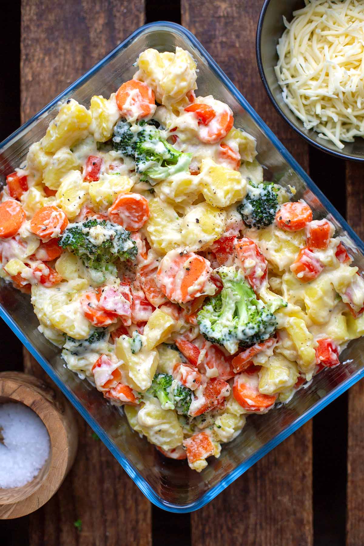 Fertiger bunter Gemüseauflauf mit Kartoffeln in einer Auflaufform. Geriebener Käse und Salz separat in einer Schüssel daneben.