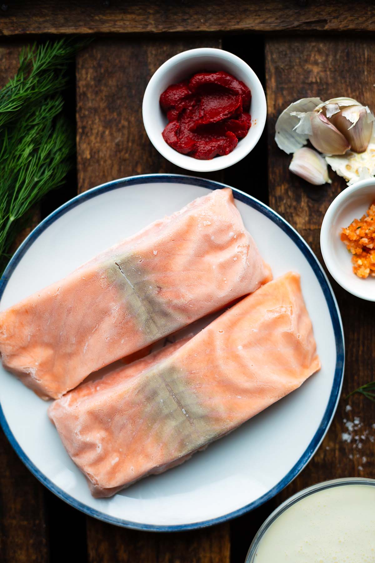 Zutaten für das Lachs-Sahne-Gratin einzeln präsentiert. Zu sehen ist: Gefrorener Lachs, Sahne, Gemüsebrühepaste, Tomatenmark. Knoblauch und Dill.