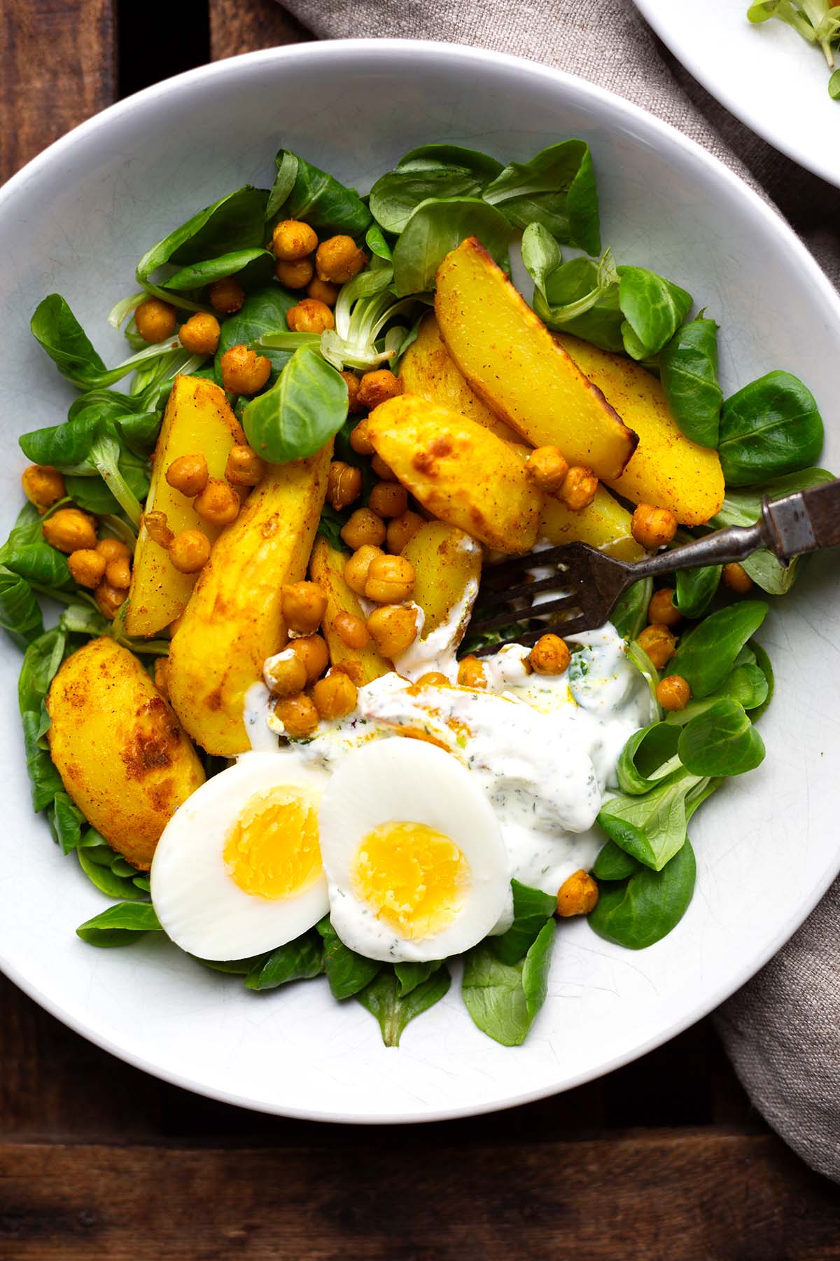 Salat Bowl mit Kichererbsen, gerösteten Kartoffel und Quark-Dressing in einer weißen Schüssel angerichtet mit einem gekochten Ei und Feldsalat.