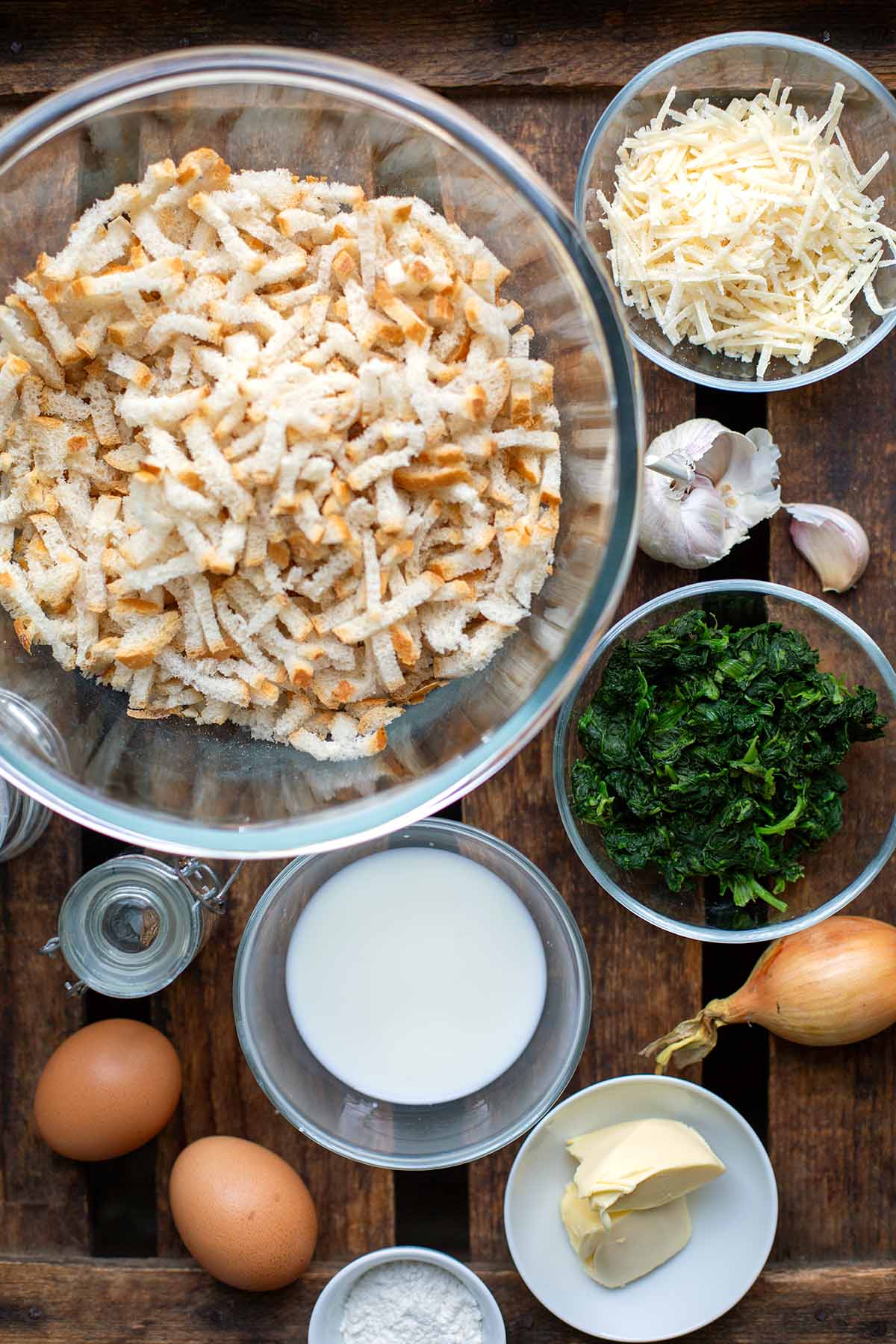 Alle Zutaten für die Spinatknödel in einzelnen Schüsseln. Zu sehen sind: klein geschnittenes Brot, Spinat, Milch, Eier, Zwiebeln und Knoblauch, Butter, Mehl und Käse.