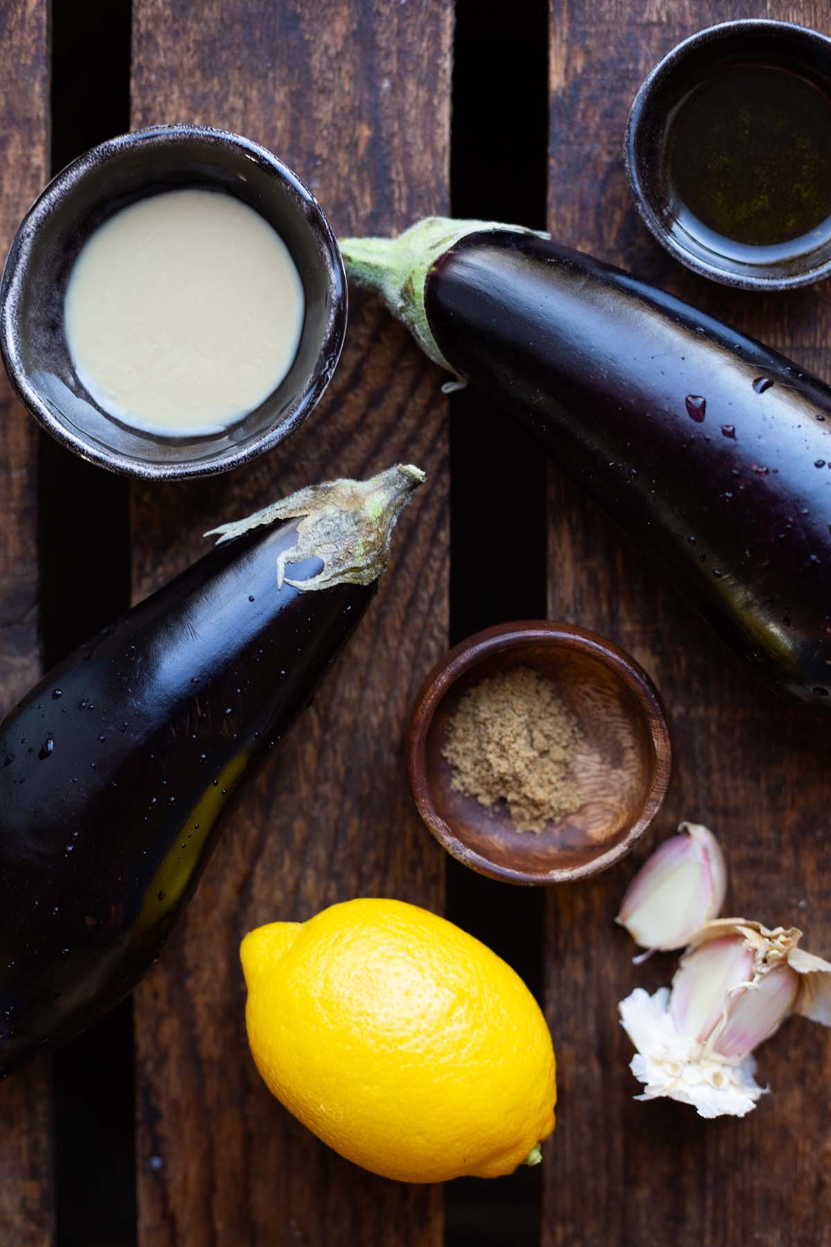 Die Zutaten für Baba Ganoush auf dunklem Holzuntergrund: Zwei Auberginen, Knoblauchzehe, Zitrone, Tahini, Kreuzkümmel und Öl in jeweils kleinen Schälchen.
