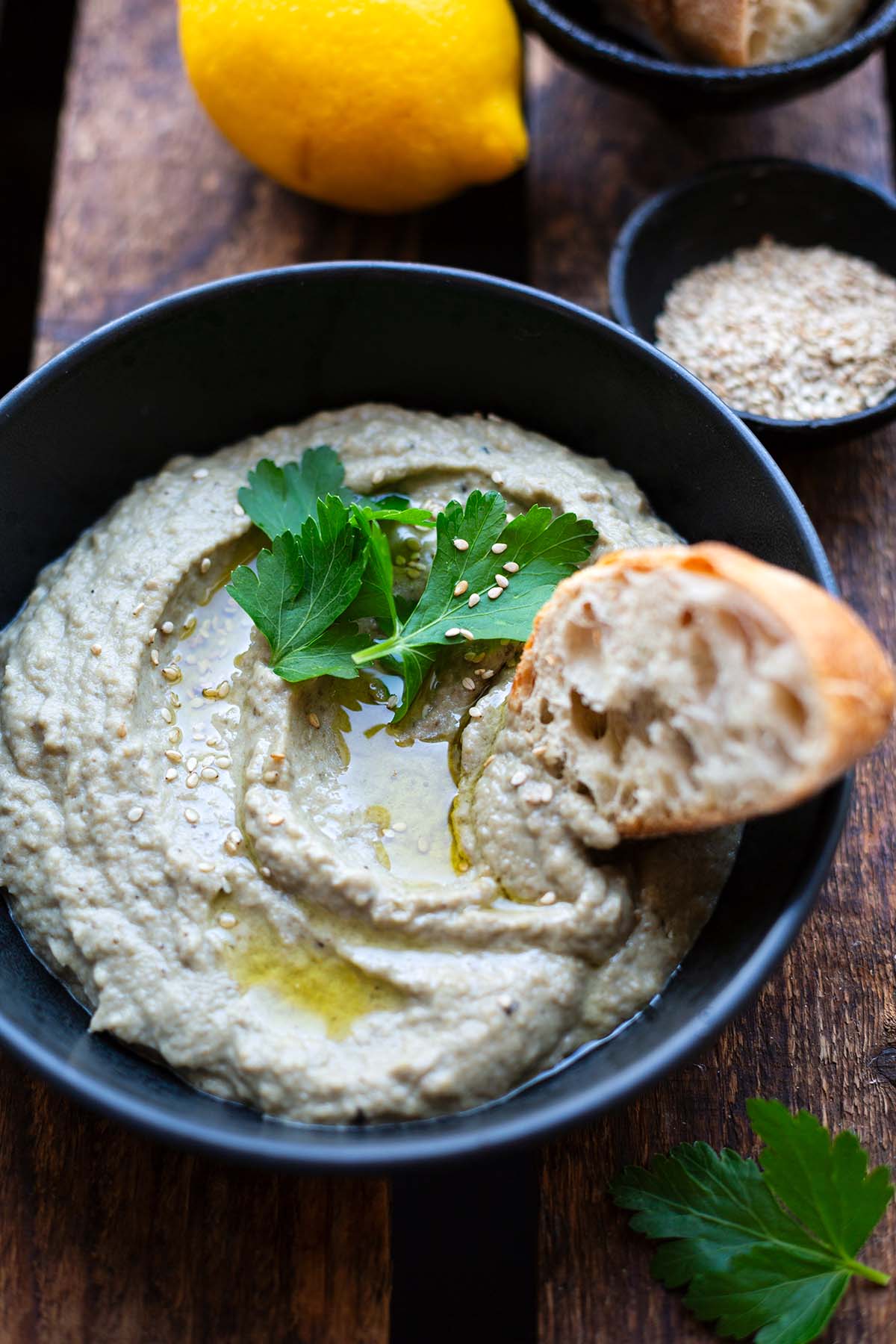 Fertiges Baba Ganoush in dunkler Schüssel auf dunklem Holzuntergrund. Garniert mit Sesam, Öl, Koriander und einer Scheibe Baguette.