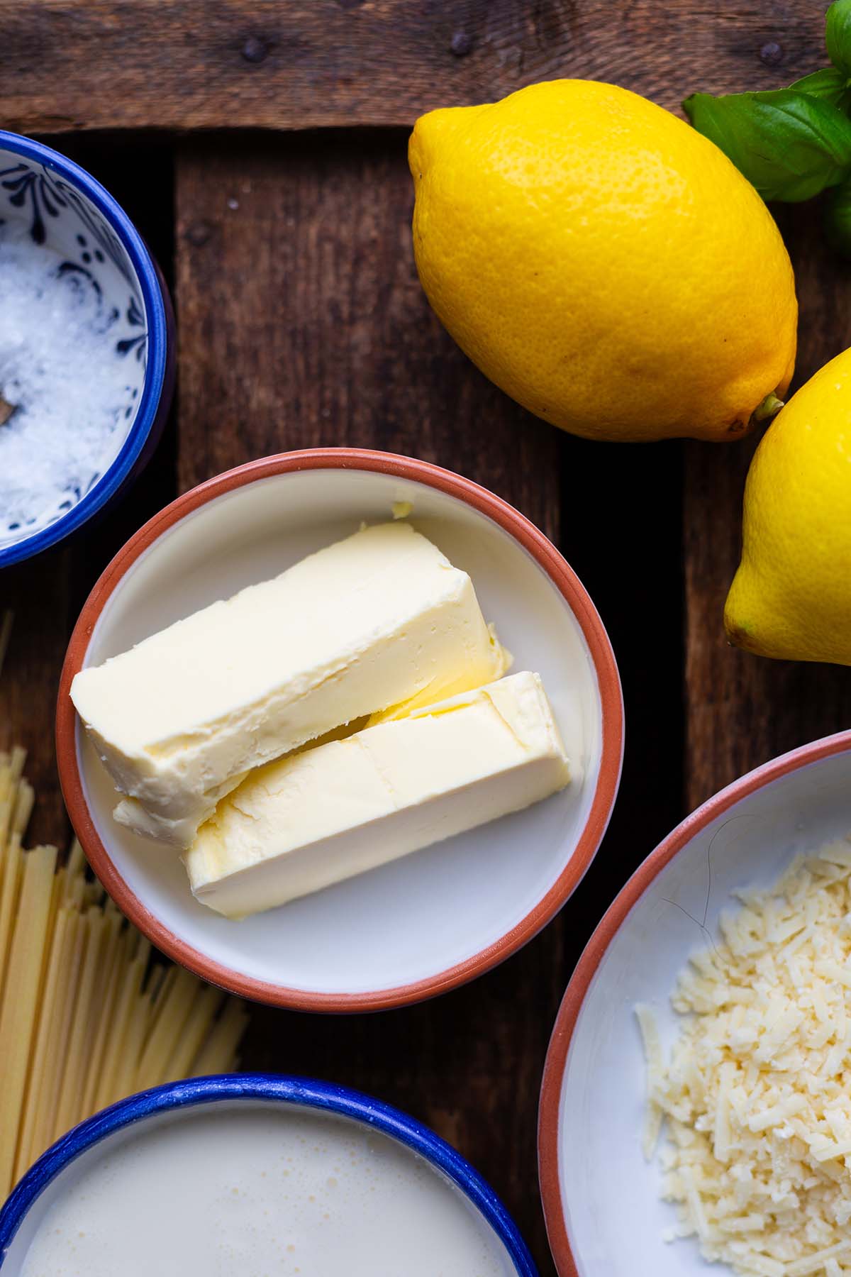Zutaten für die Pasta al limone auf dunklem Holzuntergrund. Zu sehen sind: ganze Zitronen, Butter, Sahne, Parmesan und Salz jeweils in einer Schüssel.